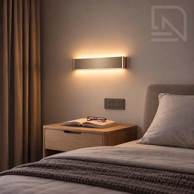 A modern rectangular wall sconce with a brushed metal finish, mounted on a bedroom wall above a wooden nightstand and lit in warm glow.