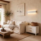 A styled living room scene featuring a cream-colored linen sofa, a rustic wooden coffee table, a white two-drawer nightstand with brass handles, and a woven basket on top. The background consists of a textured beige plaster wall with a modern linear wall sconce emitting warm light, a framed abstract print, and a tall potted tree near a window.
