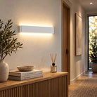 A modern rectangular wall sconce with a matte white finish, mounted on a beige wall in a warm, contemporary hallway setting.