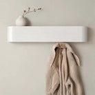 A minimalist white floating shelf mounted on a warm beige wall, styled with a small white vase holding dried stems and a beige coat hanging from the hook below.