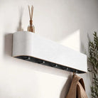 A modern wall-mounted coat rack with a textured white top and dark gray interior hooks, shown mounted on a white wall with a reed diffuser on top and a brown coat hanging from the side.