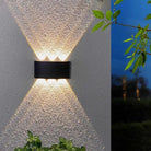 A modern black rectangular outdoor wall sconce mounted on a textured stucco wall, casting a warm light upward and downward in a V-shape. A split-screen view on the right shows the same fixture in use at dusk against a dark blue sky.