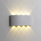 A modern rectangular wall sconce light fixture mounted on a white wall, casting warm light upward and downward in a diamond pattern.
