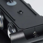 Close-up of a black metal camera accessory or mount with screws, ridges, and part of a lens visible, photographed on a light neutral background.