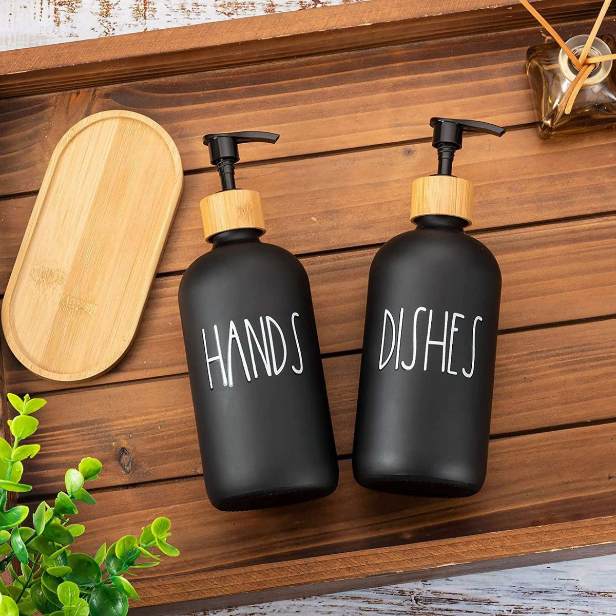 Two black soap dispensers labeled 'HANDS' and 'DISHES' on a wooden surface with a wooden tray and plant in the background.