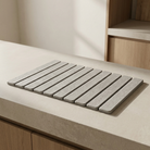 A rectangular gray slatted mat or tray with rounded corners, placed on a light beige countertop in a modern kitchen setting with wood cabinetry visible in the background.