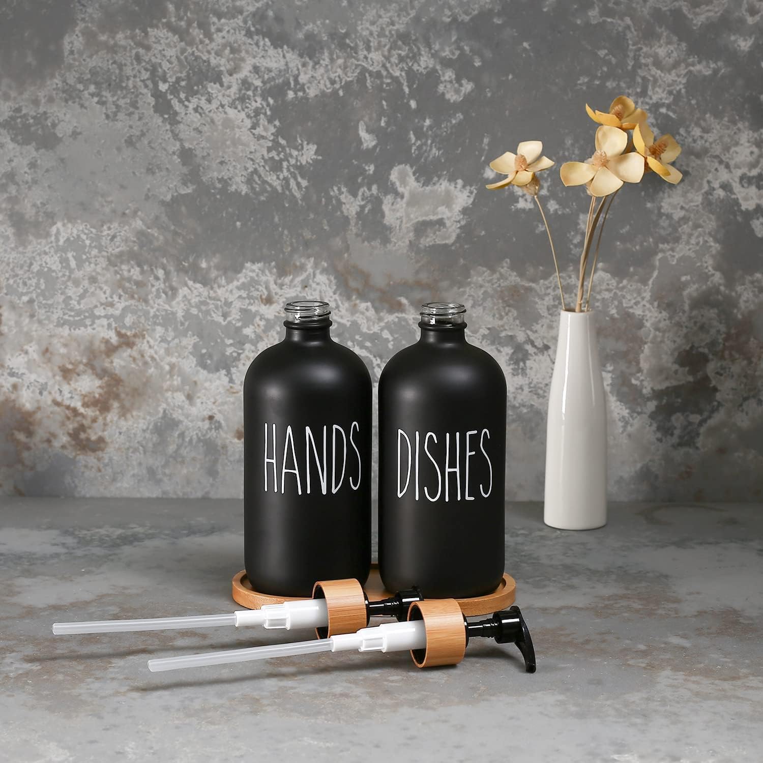 Black 'HANDS' and 'DISHES' bottles with a white vase with flowers on a textured gray background
