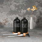 Black 'HANDS' and 'DISHES' bottles with a white vase with flowers on a textured gray background