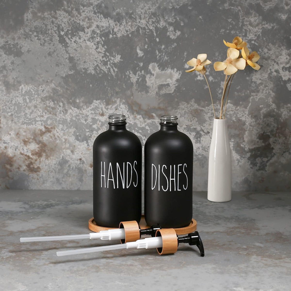 Black 'HANDS' and 'DISHES' bottles with a white vase with flowers on a textured gray background