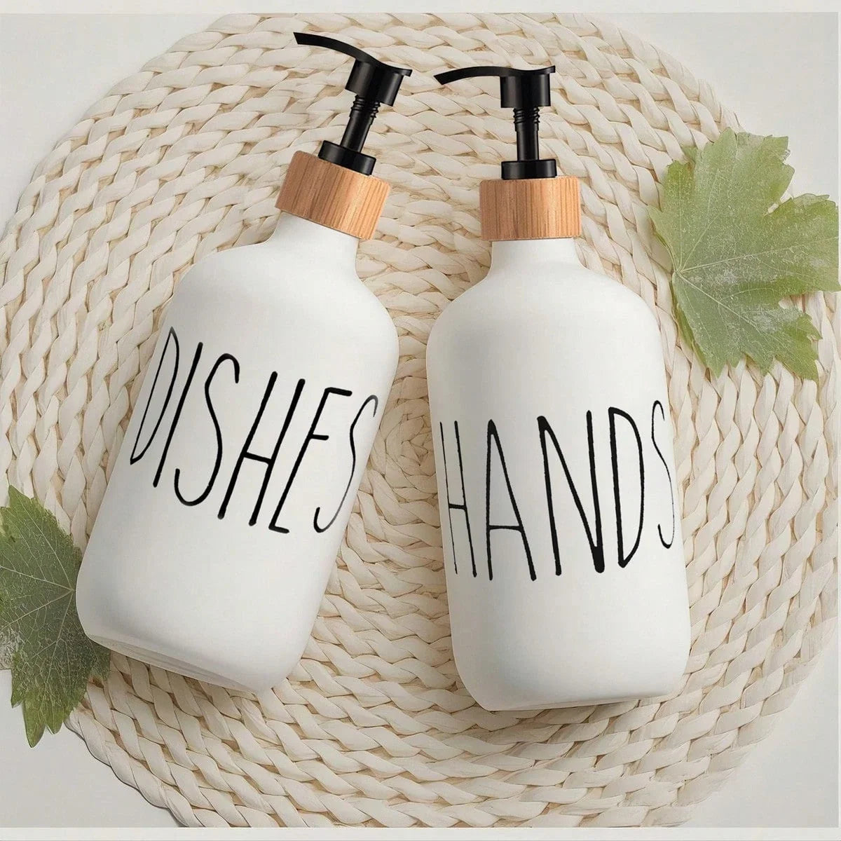 Two white soap dispensers labeled 'Dishes' and 'Hands' on a woven mat with leaves.