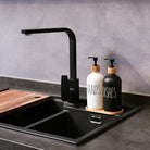 Black kitchen faucet over a sink with two soap dispensers labeled 'HANDS' and 'DISHES' on a gray countertop.